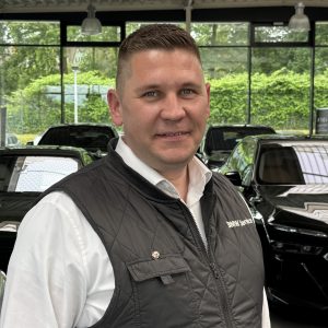 Person in schwarzer Weste vor mehreren Autos in einem Autohaus mit großen Fenstern im Hintergrund.