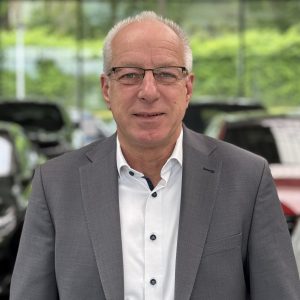 Ein Mann in Anzug und Brille steht vor unscharfen Autos in einem Autohaus.