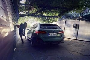 Grünes BMW-Auto lädt an einer Wallbox, Frau lehnt an einer Wand, Bäume im Hintergrund.