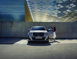 Frau steht neben einem silbernen BMW iX1 in einem modernen architektonischen Umfeld.
