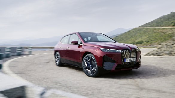 Roter BMW iX fährt auf kurviger Straße in bergiger Landschaft, bewölkter Himmel.