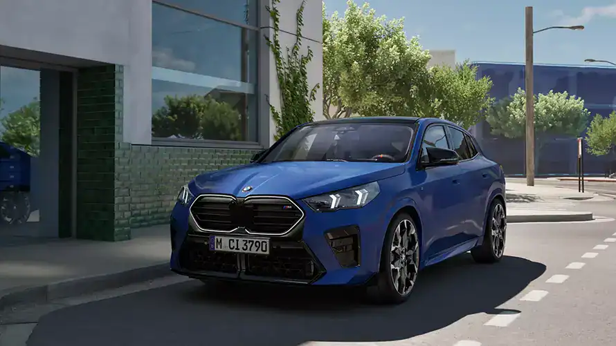 Blauer BMW X2 M auf einer Straße, umgeben von modernen Gebäuden und Bäumen bei Sonnenschein.