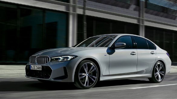 Silberner BMW 3er Limousine fährt auf einer Straße vor einem modernen Gebäude.