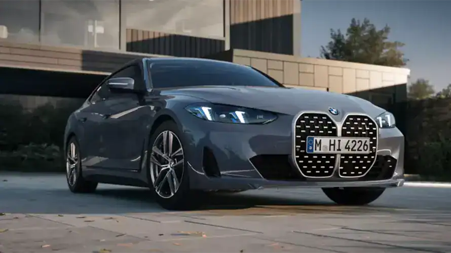 Graues BMW 4er Gran Coupé auf Parkplatz vor modernem Gebäude, beleuchtete Scheinwerfer.