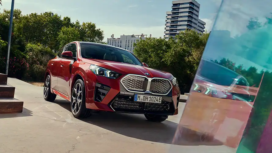 Roter BMW iX2 vor grünen Bäumen und modernem Gebäude bei Tag.