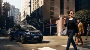 BMW i3 fährt in einer Stadt; Frau überquert Straße; moderne Gebäude im Hintergrund.