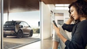 Frau sitzt im Raum, schaut auf Handy, BMW i3 lädt draußen an Ladestation.