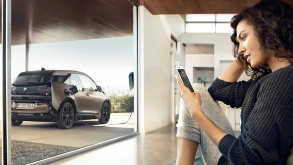 Frau sitzt drinnen mit Smartphone, draußen lädt ein BMW i3 an einer Ladestation.