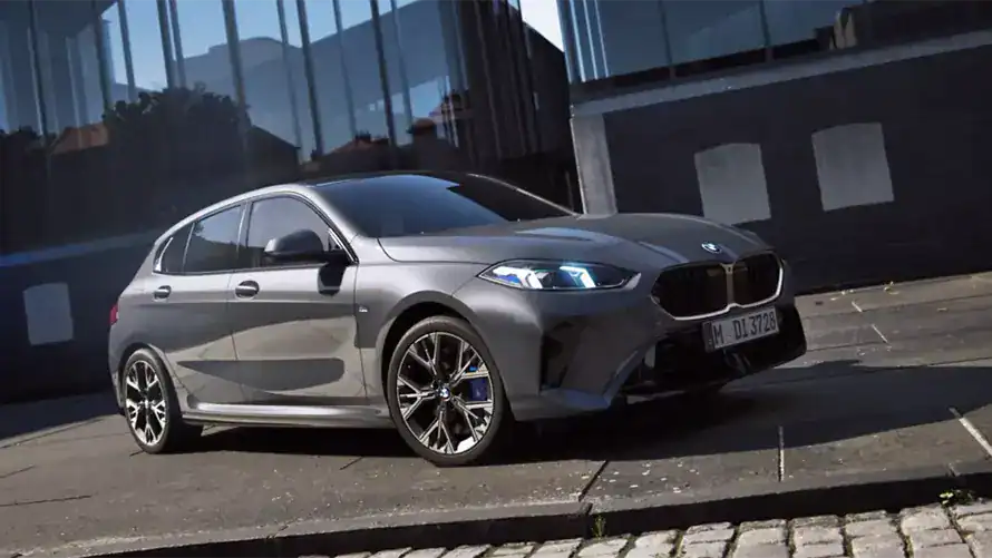 Grauer BMW 1er parkt vor einem modernen Gebäude auf einem gepflasterten Gehweg.