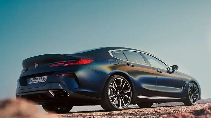 BMW 8er Gran Coupé in dunkler Farbe, seitliche Heckansicht, auf einer Straße bei klarem Himmel.