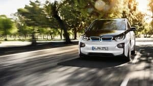BMW i3 fährt auf sonniger Straße, umgeben von grünen Bäumen.