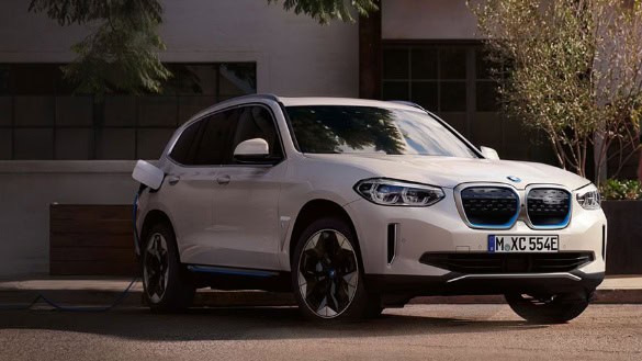 Weißer BMW-SUV parkt im Freien, angeschlossen an Ladestation, Bäume im Hintergrund.