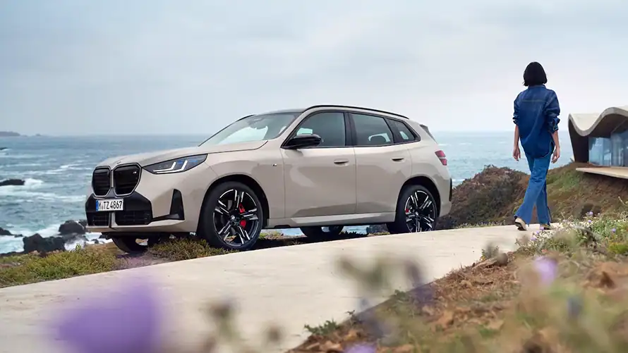 BMW X3 M50 in Beige an einer Küstenstraße, Frau in Jeansjacke und -hose geht weg, Meer im Hintergrund.