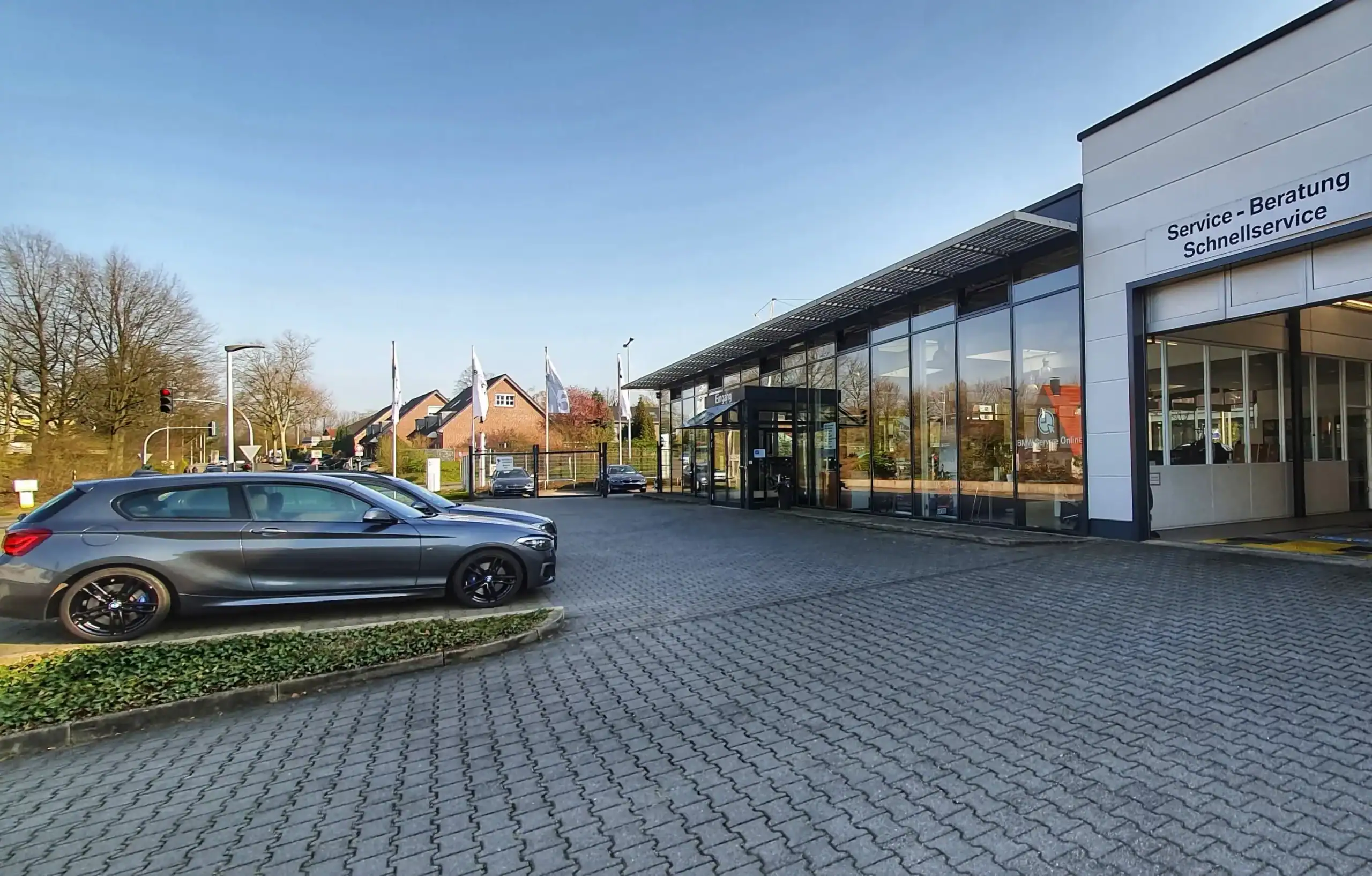 BMW Autohaus mit grauem Auto im Vordergrund und verglastem Gebäude im Hintergrund, blauer Himmel.