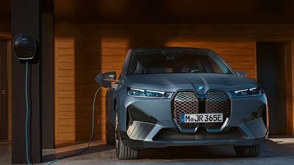 Graues BMW iX M60 Elektroauto lädt an einer Ladestation vor einem Haus mit Holzwänden.