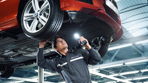 Mechaniker inspiziert rotes Auto auf Hebebühne mit Taschenlampe in BMW Autohaus Mülheim.