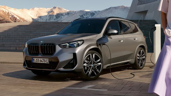 Grauer BMW SUV an Ladestation im Freien, Berge im Hintergrund.