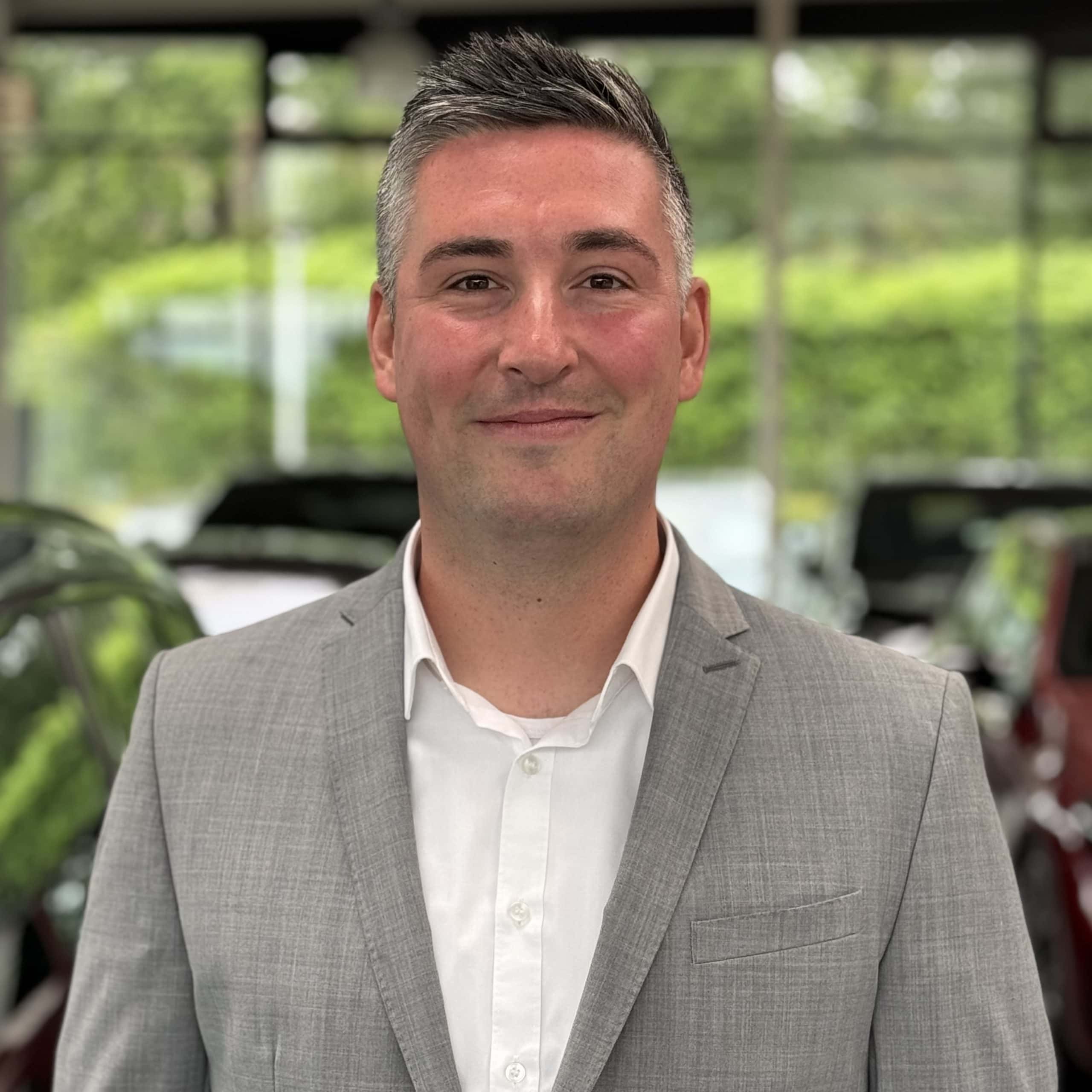 Mann im Anzug lächelt in Kamera; Hintergrund unscharf, möglicherweise BMW Autohaus Mülheim.