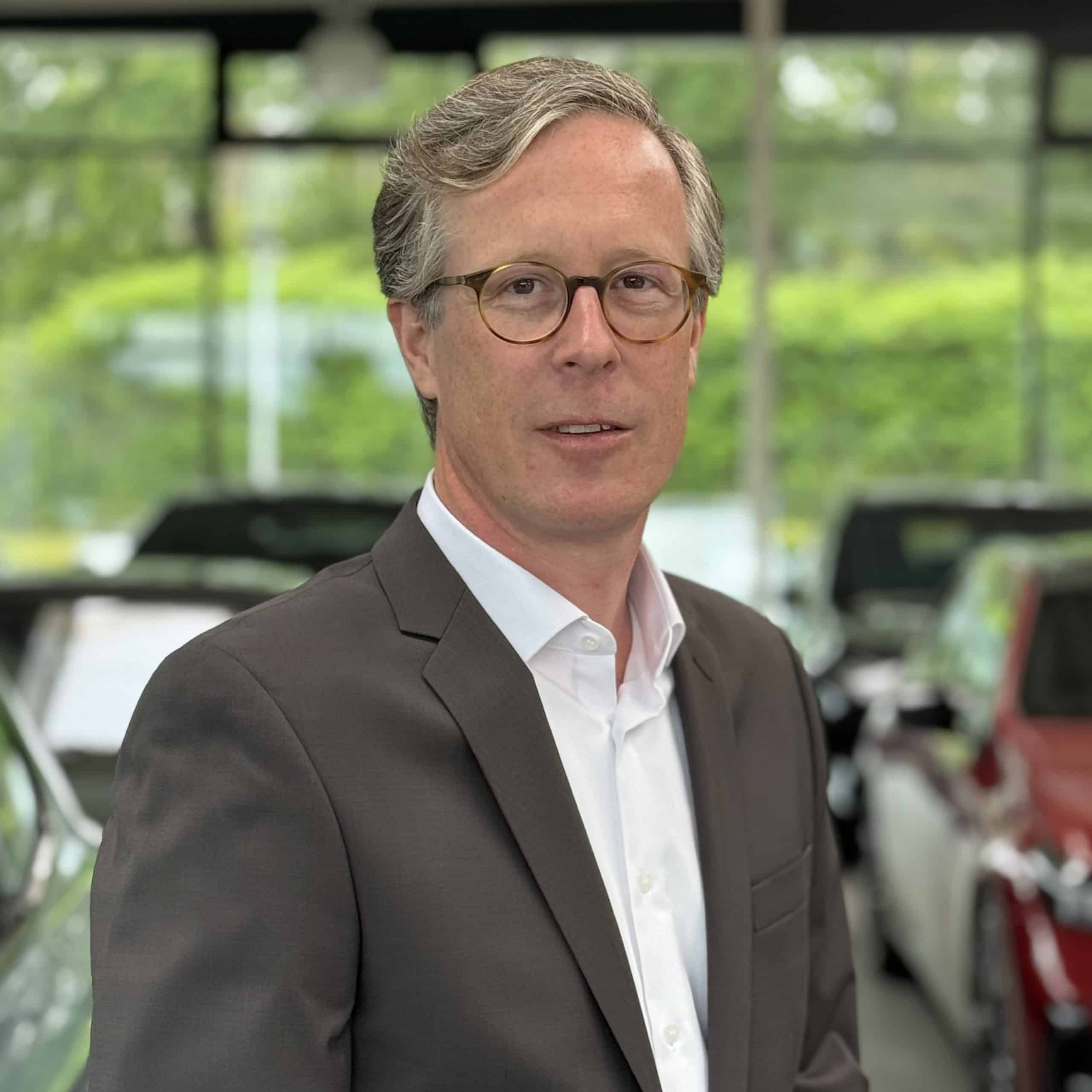 Mann im Anzug vor einem Auto im BMW Autohaus Mülheim, lächelnd, mit Brille und Krawatte.